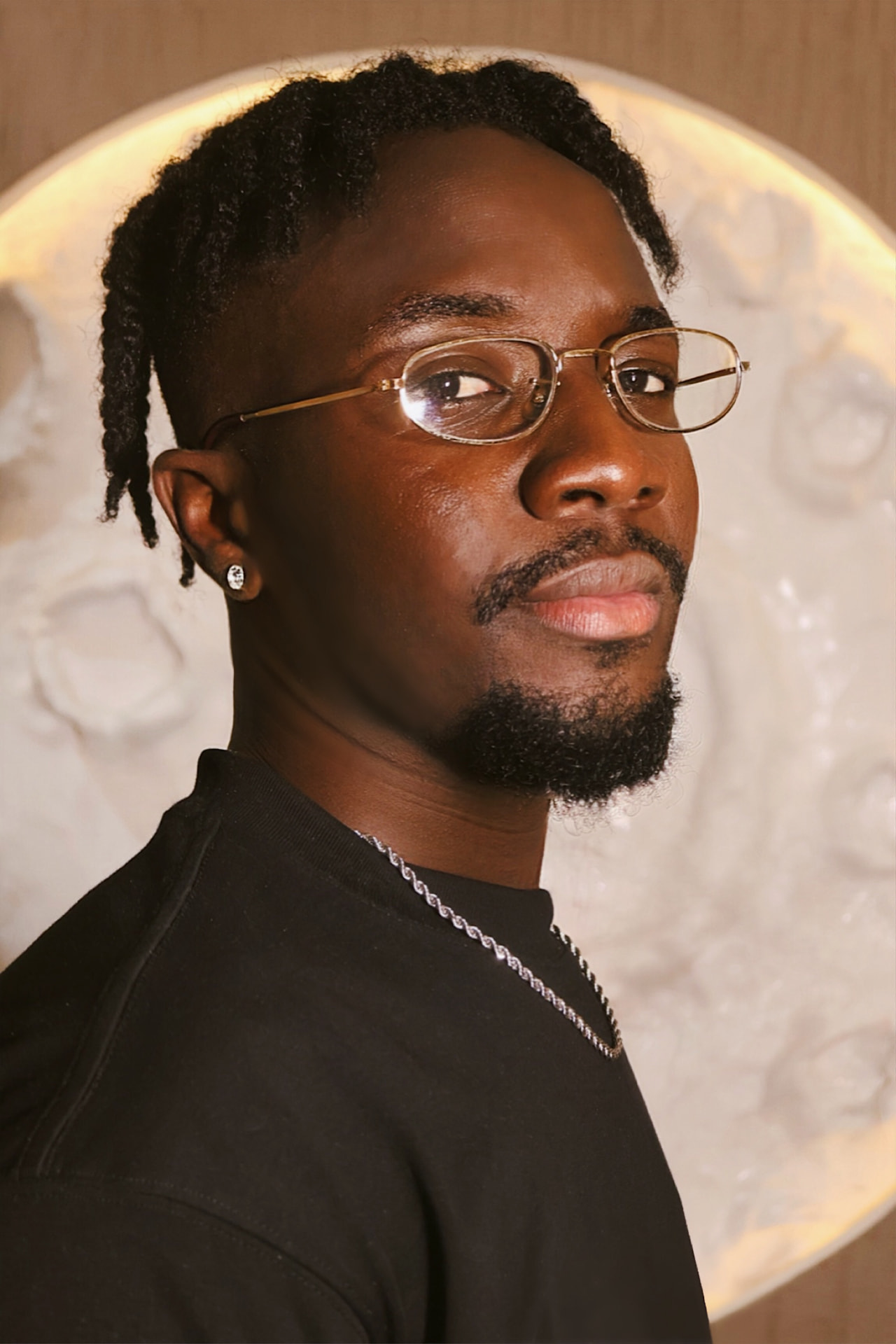 Broni in a black shirt and wire-rimmed glasses against a warm backlit wall